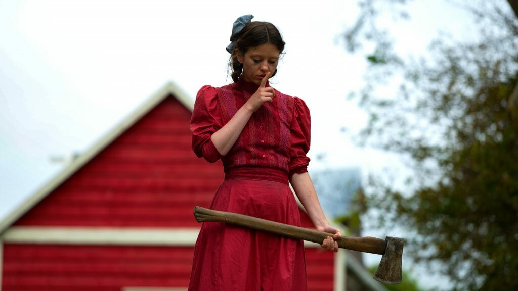 Mia Goth stars as Pearl wearing a bright red dress in front of a red barn telling her next victim to shush with one hand and holding a large axe in the other in the A24 horror film.