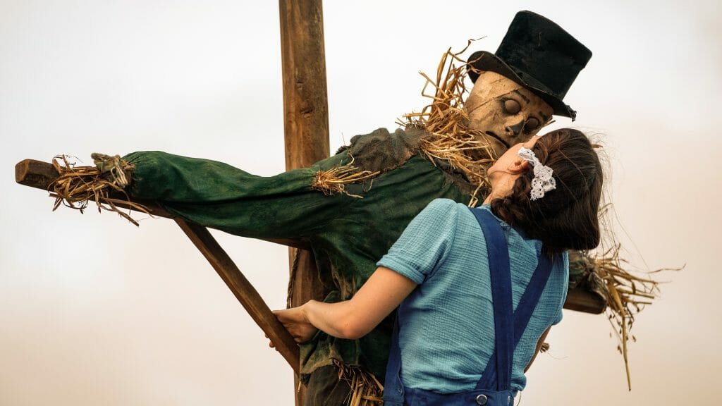 Mia Goth stars as Pearl in bright blue overalls climbing an old scarecrow wearing a top hat and giving it a kiss on the lips in the A24 horror film from Ti West. 