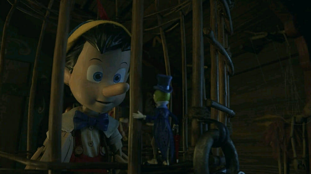 Pinocchio voiced by Benjamin Evan Ainsworth talks to Jiminy Cricket voiced by Joseph Gordon-Levitt while trapped in a metal cage in the 2022 live-action film on Disney+.