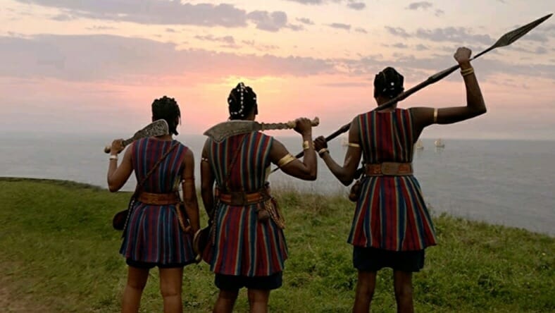 Three Agojie female warriors holding battle swords and spears overlook a beautiful sunset over the ocean next to the Dahomey empire in THE WOMAN KING. 