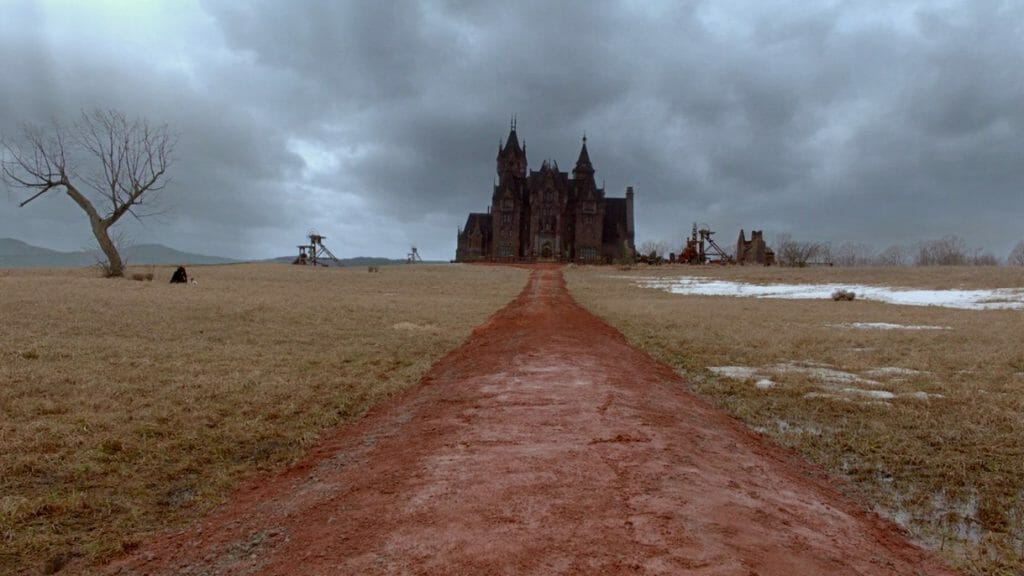 A red clay road leads up to the haunted Sharpe family mansion of Crimson Peak under a dark gray sky as seen in the Gothic Romance directed by Guillermo del Toro. 
