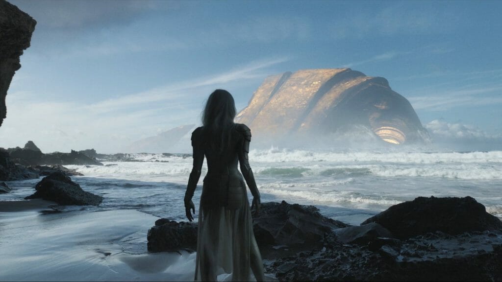 Thena played by Angelina Jolie stands on a rocky beach shore and stares at the giant head of Tiamut the Golden Celestial rising from the Earth's crust in the Ocean in ETERNALS directed by Chloé Zhao.
