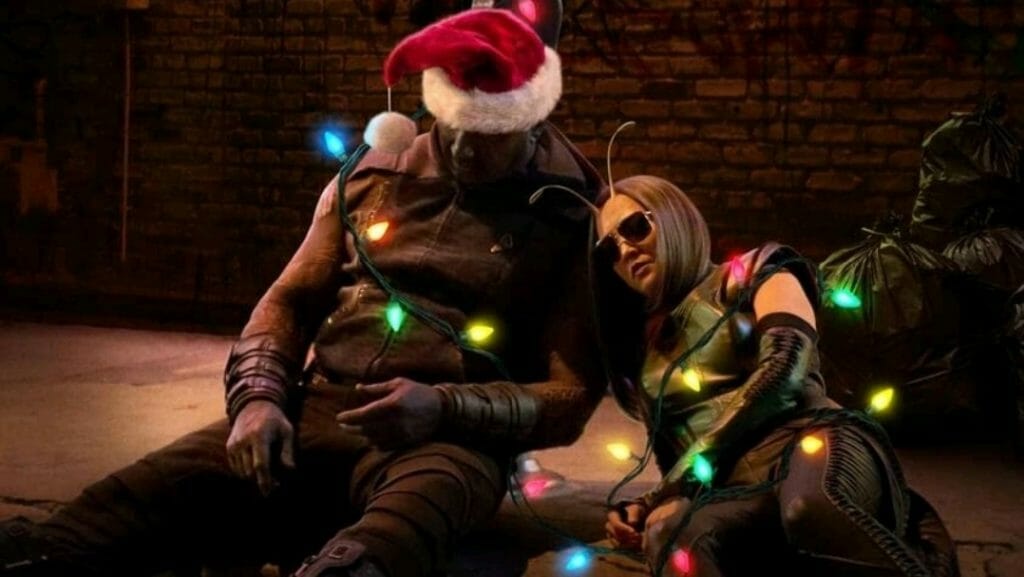 Drax wearing a Santa Claus hat and Mantis wearing Sunglasses lay wasted on the curb of a Hollywood sidewalk covered in Christmas lights from THE GUARDIANS OF THE GALAXY HOLIDAY SPECIAL.