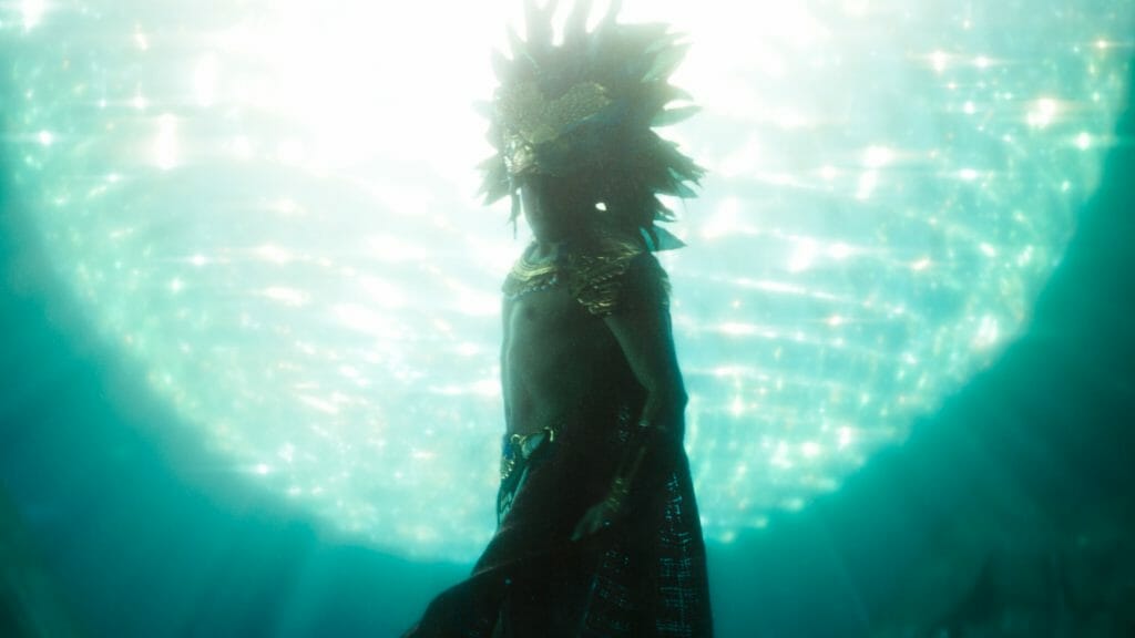 Tenoch Huerta as Namor wears his Kukulkan Mayan head piece and royal robes while swimming over the ancient throne of the underwater city Talokan in the MCU film BLACK PANTHER: WAKANDA FOREVER. 