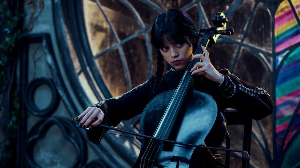 Jenna Ortega as Wednesday Addams plays the Cello with an emotionless stare in front of her stain glassed window from her Nevermore Academy dorm room in the Netflix comedy series.