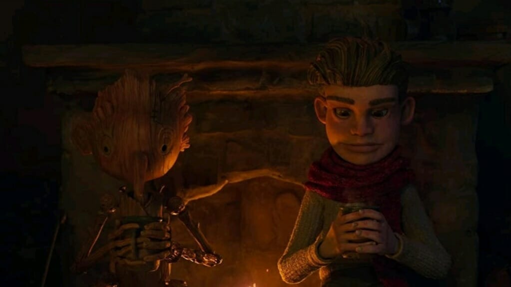 Pinocchio voiced by Gregory Mann and the young Italian boy Candlewick voiced by Finn Wolfhard sit down and share hot chocolate by a chimney fire in the stop-motion animated film GUILLERMO DEL TORO'S PINOCCHIO on Netflix.