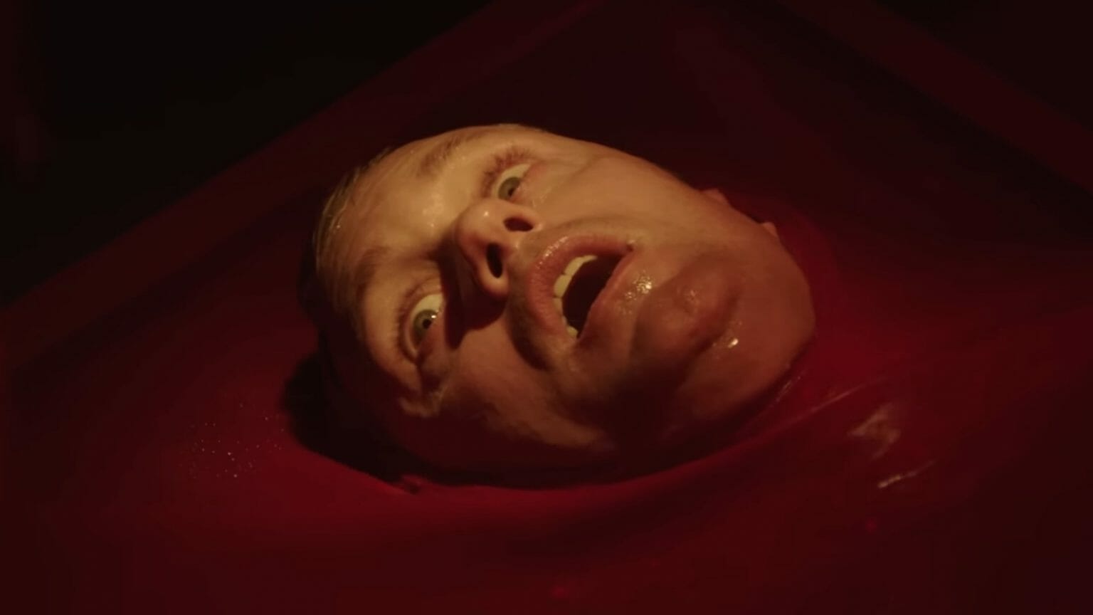 The face of a newborn Alexander Skarsgård clone wakes up shocked in fear from a red rubber looking vessel in the body horror film INFINITY POOL directed by Brandon Cronenberg.