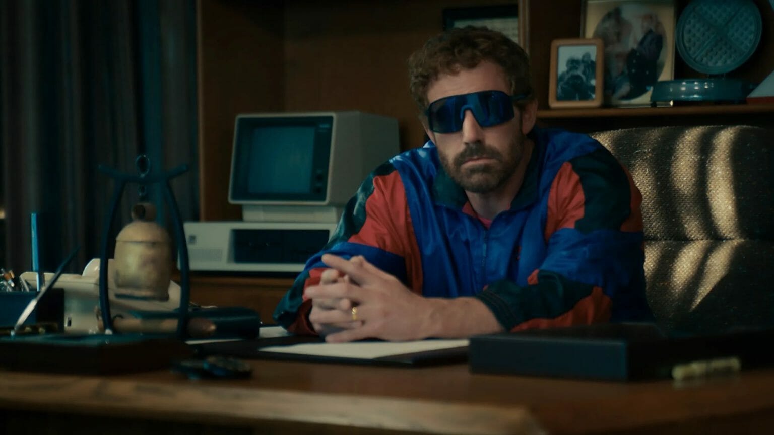 Ben Affleck stars as Nike CEO Phil Night wearing large blue goofy sunglasses and a matching tracksuit while sitting in his corporate office in the movie AIR from Amazon Studios.