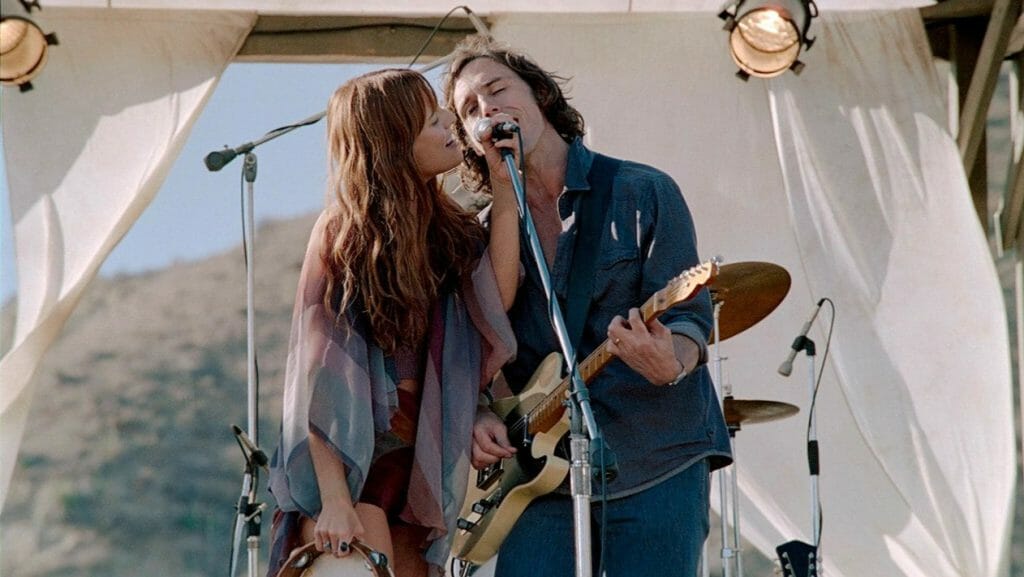 Riley Keough as lead singer Daisy Jones holding a tambourine and Sam Claflin as guitarist Billy Dunne singer together passionately on stage in the Amazon Prime Video musical adaptation of DAISY JONES & THE SIX.