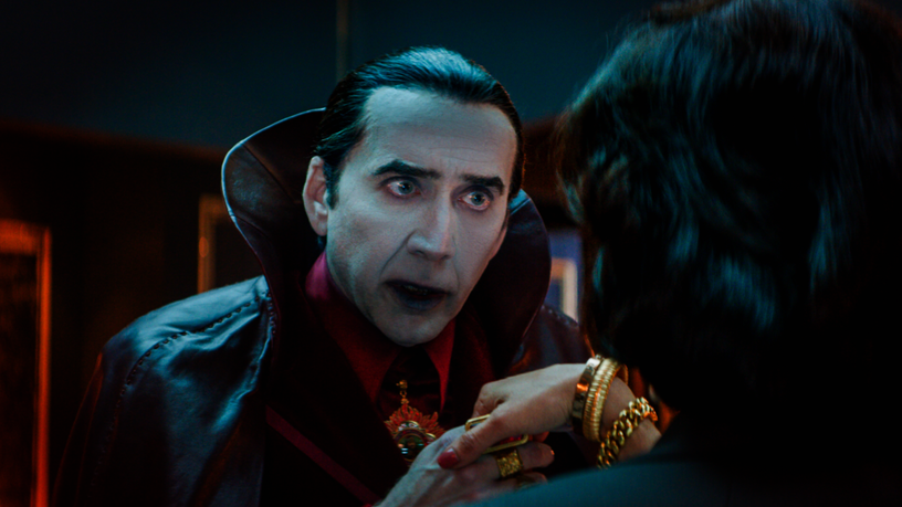 Nicolas Cage stars as Dracula with white make-up in the classic red and black cloak greeting a woman by kissing her hand in the 2023 horror comedy film RENFIELD.