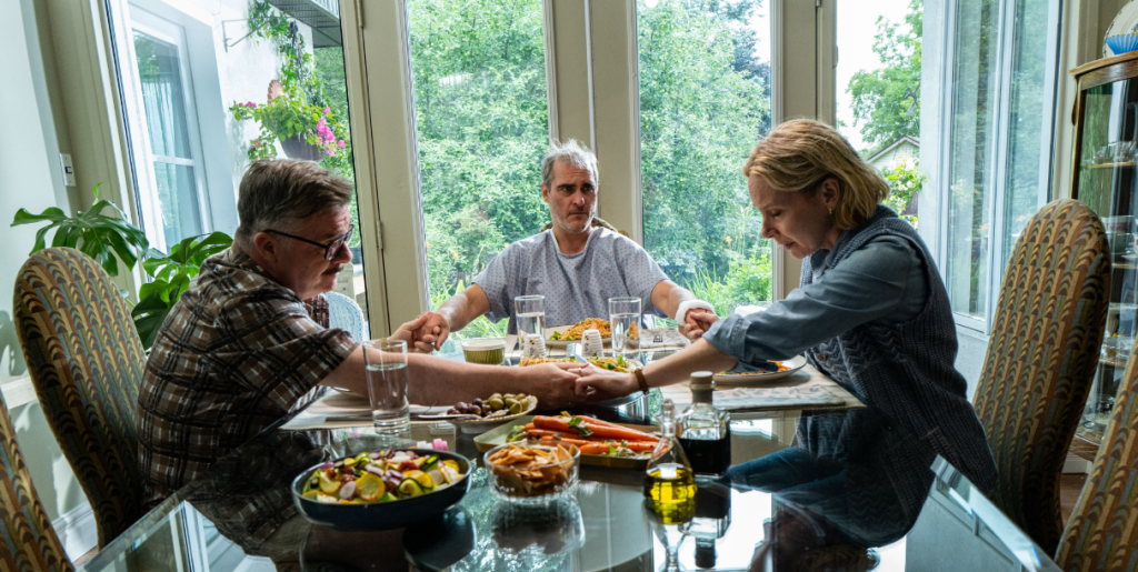Joaquin Phoenix as Beau sits looking confused at a fancy dinner table and holds the hands of Nathan Lane as father Roger and Amy Ryan as mother Grace while they save a prayer in the A24 black comedy horror film BEAU IS AFRAID.