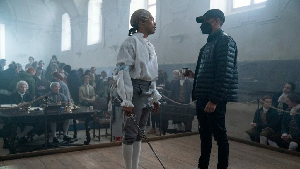 Lead actor Kelvin Harrison Jr. in costume as Joseph Bologne holds a sword and rehearses a fencing scene with director Stephen Williams in front a crowd of actors in period costumes on the set of the biopic film CHEVALIER.