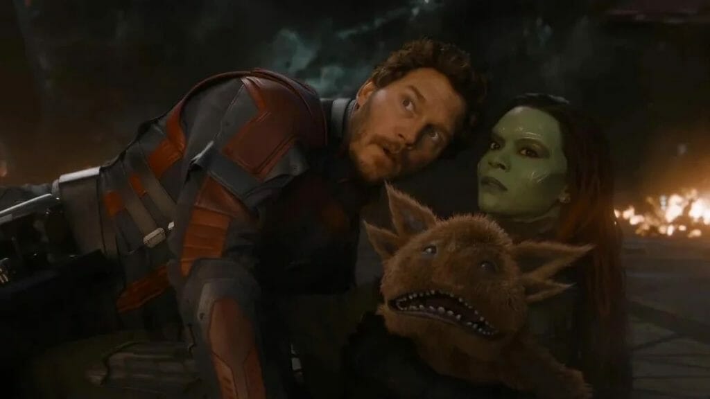 Chris Pratt as Star-Lord and Zoe Saldana as Gamora look at an explosion while getting up from a fiery battlefield together with a bizarre looking brown alien critter cuddles up to them in GUARDIANS OF THE GALAXY VOL. 3