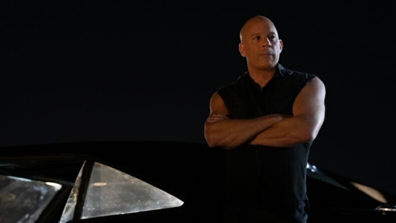 Vin Diesel stars as Dominic Toretto wearing a black vest and posing with his arms crossed while leaning on his new black charger in FAST X.