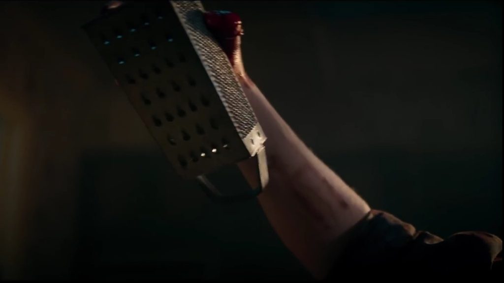 The hand of a deadite raises a large metal cheese grater in the air preparing to attack in EVIL DEAD RISE. 