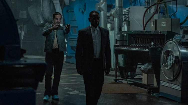 Lalo Salamanca holds Gus Fring at gun point and walks him into an empty dark laboratory in BETTER CALL SAUL Season 6.