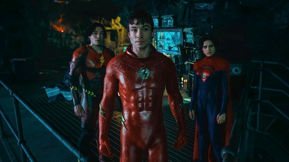 Ezra Miller as two versions of Barry Allen, one younger with long hair and the other older with short hair, in two different colored Flash costumes stand next to Sasha Calle as Supergirl in the Batcave in THE FLASH. 