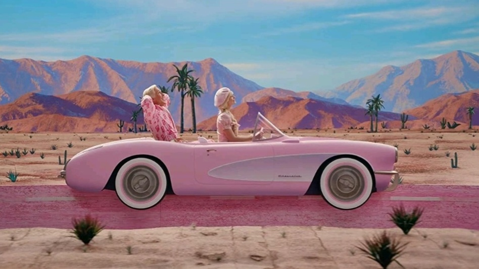 Barbie played by Margot Robbie drives a pink convertible Corvette car across the desert while Ken played by Ryan Gosling sits in the back seat in a relaxed position with his arms behind his head.