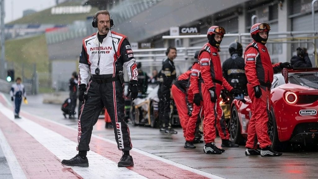 David Harbour stars as coach Jack Salter wearing a Nissan uniform on the race track standing by his pit crew in the 2023 GRAN TURISMO movie. 