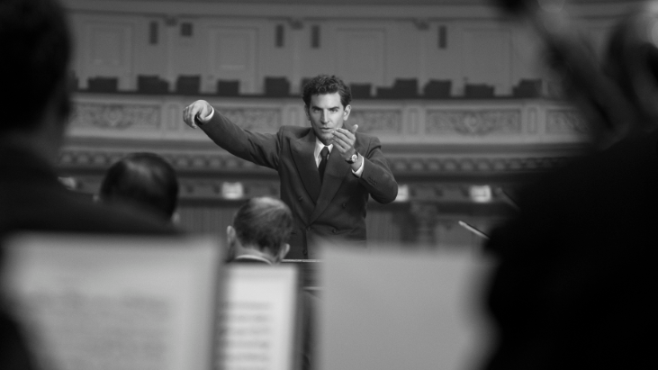 Bradley Cooper as a young Leonard Bernstein conducting an orchestra at the New York Philharmonic in a black-and-white shot from the movie MAESTRO. 