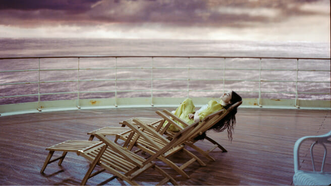 Emma Stone stars as Bella Baxter laying on the deck of ship in a beautiful yellow dress in front of an open violet-colored ocean in a cinematic shot from the movie POOR THINGS. 