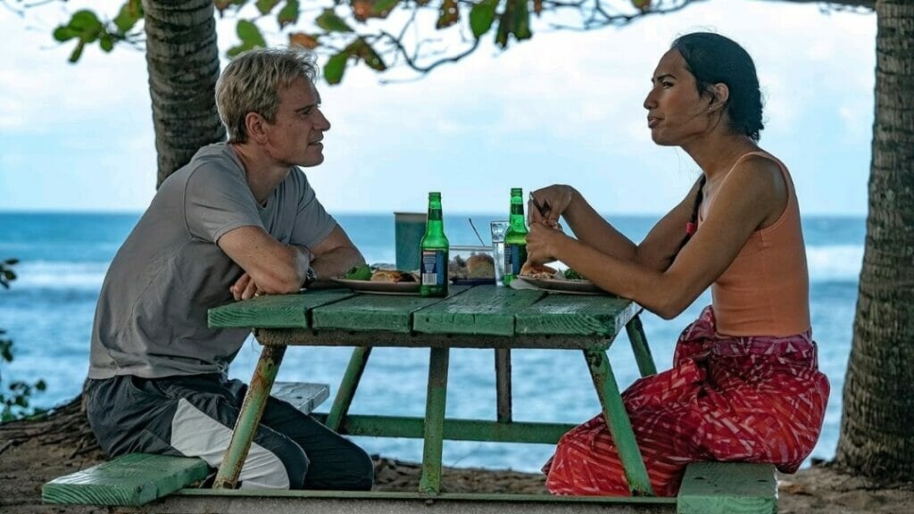 Michael Fassbender as soccer coach Thomas Rongen sits down for lunch with Jaiyah, a fa'afafine member of the American Samoa national team played by Kaimana in the movie NEXT GOAL WINS.