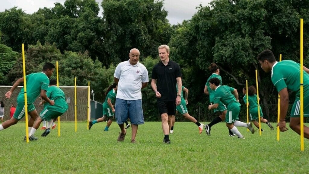 Coach Thomas Rongen played by Michael Fassbender walks down a green field with team owner Tavita played Oscar Kightley as the American Samoa national soccer team trains around them in the sports comedy NEXT GOAL WINS.
