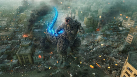 Godzilla lets out a loud roar while standing the middle of destroyed buildings in Tokyo with his dorsal plates glowing bright blue with atomic energy in the movie GODZILLA MINUS ONE.