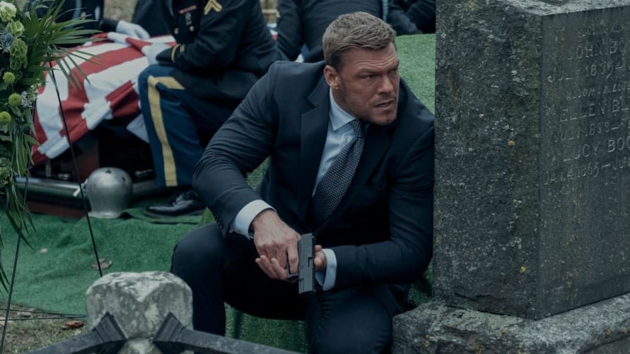 Alan Ritchson stars as Jack Reacher taking cover from enemy fire behind a gravestone in an intense action sequence from REACHER Season 2 coming to Amazon Prime Video in December 2023.