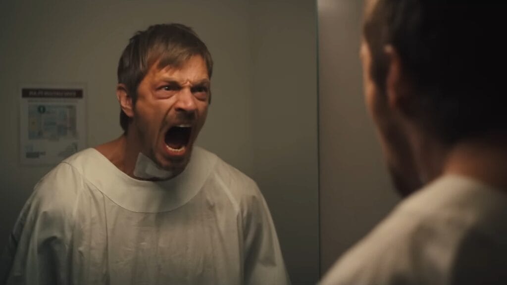 Joel Kinnaman discovers that he can't speak as he yells at his reflection intensely in a hospital mirror with a bandage on his throat in the action movie SILENT NIGHT directed by John Woo. 