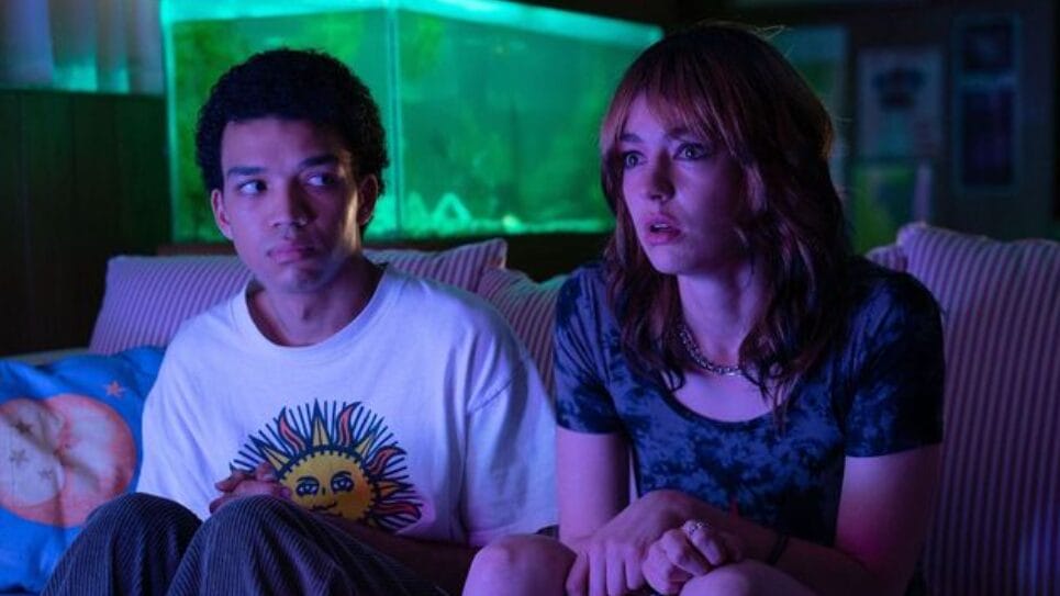 Justice Smith and Brigette Lundy-Paine look entranced as they sit together on a couch in front of a bright television in the A24 horror movie I SAW THE TV GLOW.