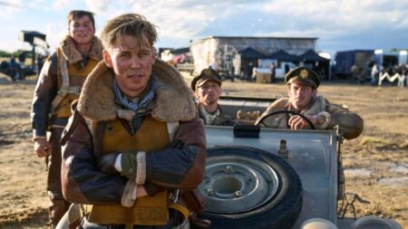 Austin Butler sits on the hood of a car in his airmen uniform with Callum Turner and Edward Ashley sitting in the front seat as they all pose together on an airfield for the MASTERS OF THE AIR miniseries on AppleTV+.