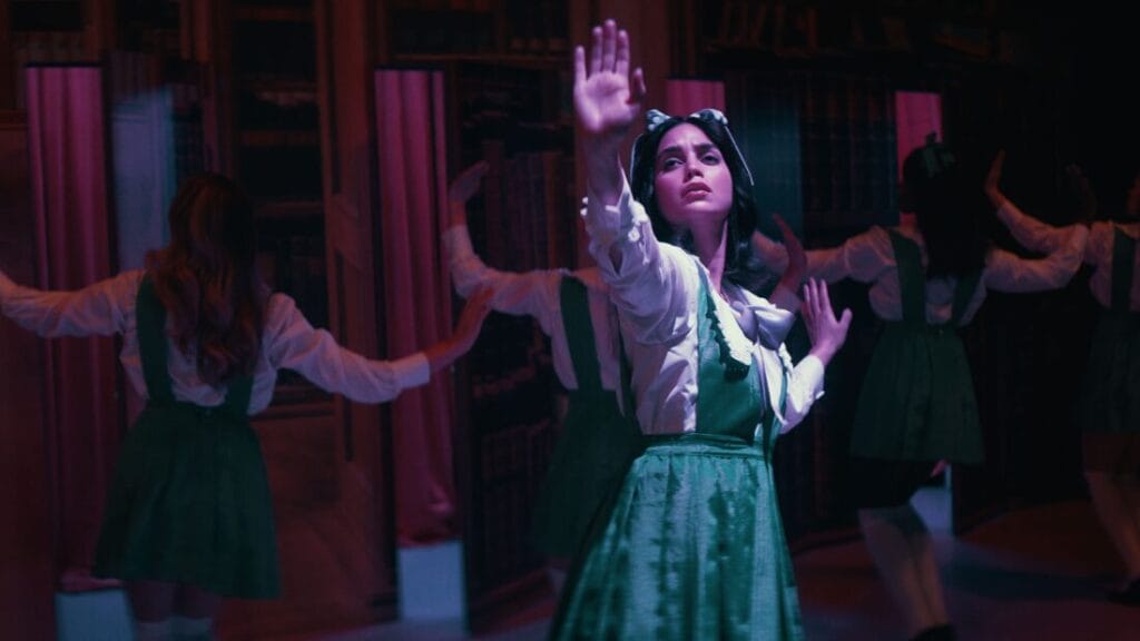 Melissa Barrera sings and poses in the spotlight in a green school girls uniform with ensemble dancers around her on stage in the horror romantic comedy YOUR MONSTER. 