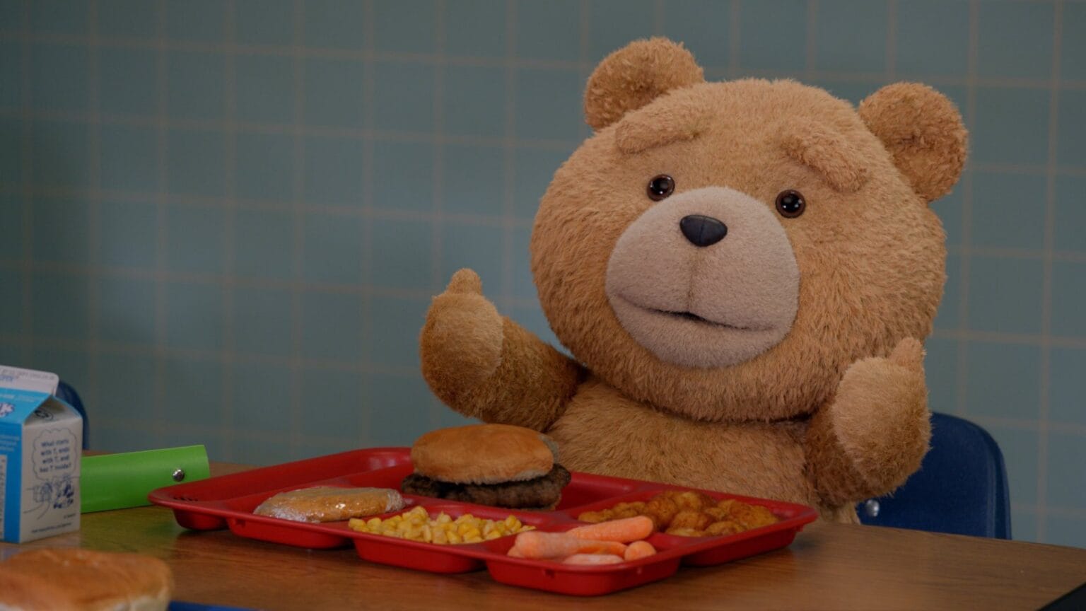 Ted voiced by Seth MacFarlane gives two thumbs up while he sits at a high school lunch table with a tray of food in the TED prequel series streaming on Peacock.