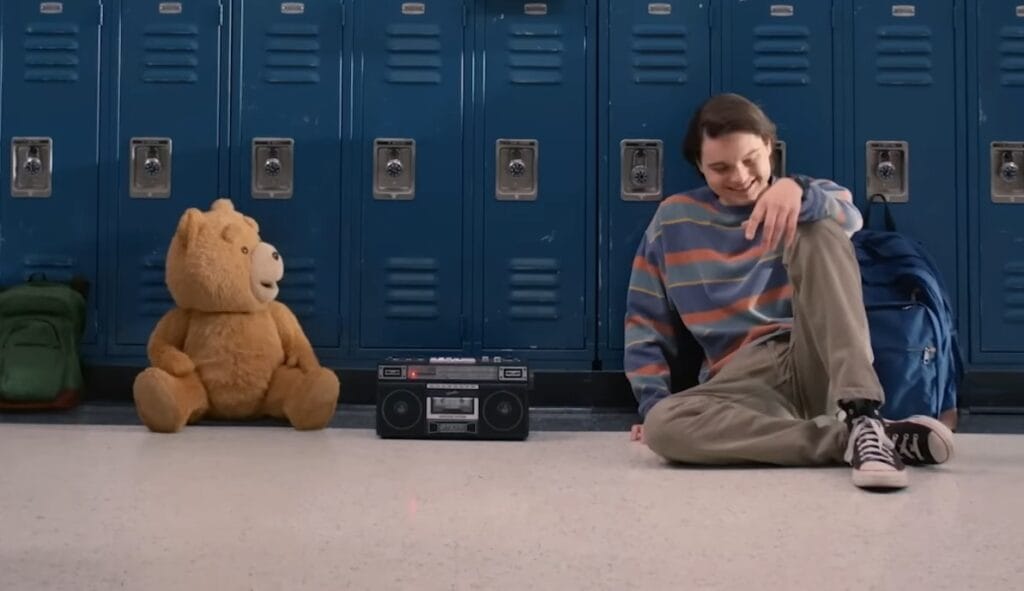 The young version of Johnny Bennett sits next to his best friend Ted the talking teddy bear on the floor of his high school in front of the lockers in the TED prequel series streaming on Peacock. 