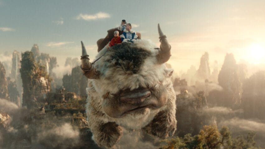 Aang, Katara, and Sokka ride on top of Appa the friendly sky bison as they fly over a beautiful canyon in the live-action AVATAR: THE LAST AIRBENDER series on Netflix. 