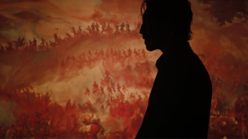 A dark silhouette of actor Dev Patel walking down a hallway in front of a large painted mural of the Hindu deity Hanuman in the action revenge thriller MONKEY MAN.  