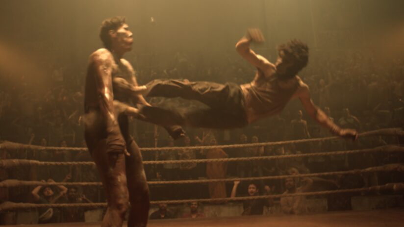 Dev Patel wearing a gorilla mask dropkicks his opponent in mid-air during a brawl in an underground fight club in the action revenge thriller movie MONKEY MAN. 