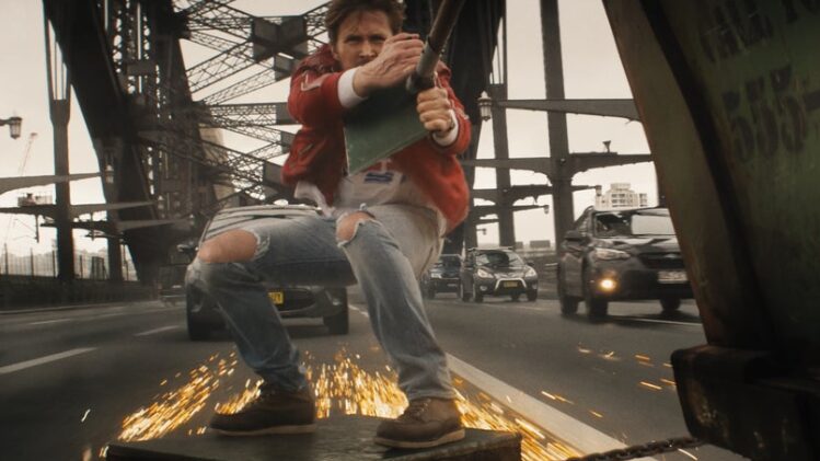 Ryan Gosling as the darring stuntman Colt Seavers hangs onto the back of a truck while skidding on a metal plate on a bridge highway during one of the action scenes in THE FALL GUY.  