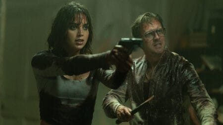 Melissa Barrera holds up a gun next to Dan Stevens holding up a wooden stake as the two are covered in blood and mud in the vampire horror film ABIGAIL.