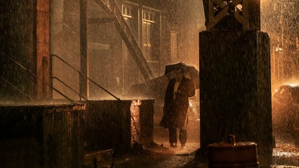 Oswald "Oz" Cobb played by Colin Farrell holds an umbrella over his head as he walks down a moody alley in Gotham City during the pouring rain in the HBO original series THE PENGUIN. 