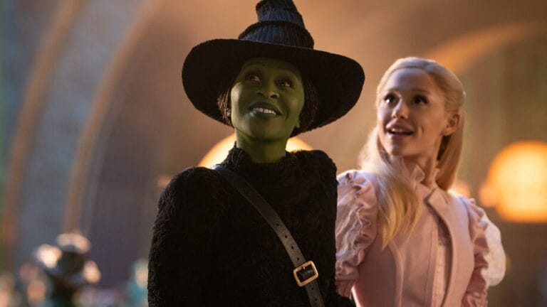 Cynthia Erivo as Elphaba Thropp wearing the iconic black witch hat locks arms with Glinda Upland played by Ariana Grande in a lavish pink dress as the two smile and explore the Emerald City together for the first time in the movie adaptation of WICKED.