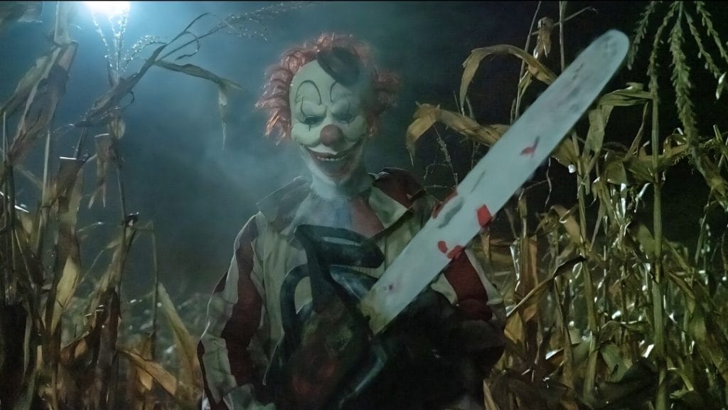 ‘Clown in a Cornfield’ Review – Meta Slasher Revitalizes the Genre with Big Laughs and Gnarly Kills Frendo the Clown holds up a large chainsaw as he runs through a thick cornfield in the 2025 meta slasher movie CLOWN IN A CORNFIELD.