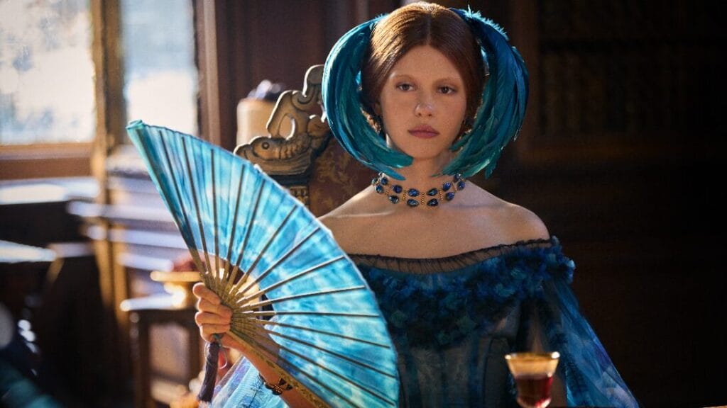 Mia Goth stars as Elizabeth Lavenza wearing a lavish 19th-century blue dress and head piece as she cools herself with a large matching blue folding fan in Guillermo del Toro's FRANKENSTEIN.