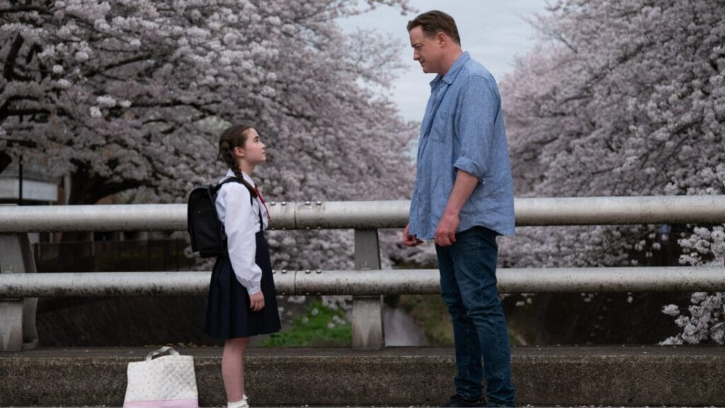 A young girl wearing a tidy school uniform awkwardly stares up at Brendan Fraser in front of a vibrant garden of cherry blossom trees in Tokyo, Japan in the dramatic comedy film RENTAL FAMILY.