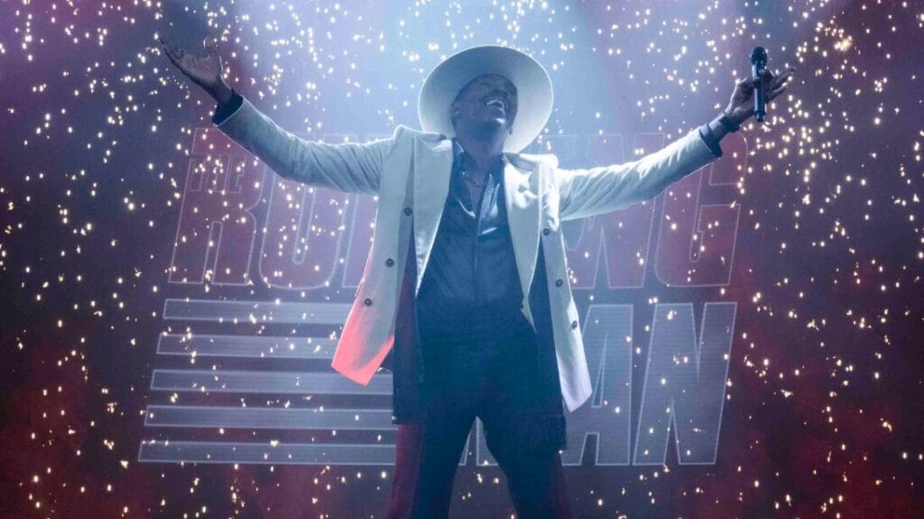 Colman Domingo stuns as TV host Bobby T. in a white suit and fedora while he stands in the middle of The Running Man stage as red and white confetti falls all around him. 