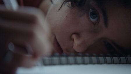 A creepy close-up shot of Tatiana Maslany's face as she holds it down to a table while drawing uncontrollably with a black pencil in the horror movie KEEPER.