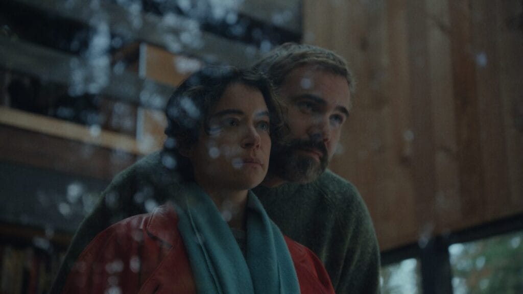 Rossif Sutherland embraces Tatiana Maslany with a romantic hug from behind as the couple stare out of a large glass window in their remote house in the woods in the horror movie KEEPER.