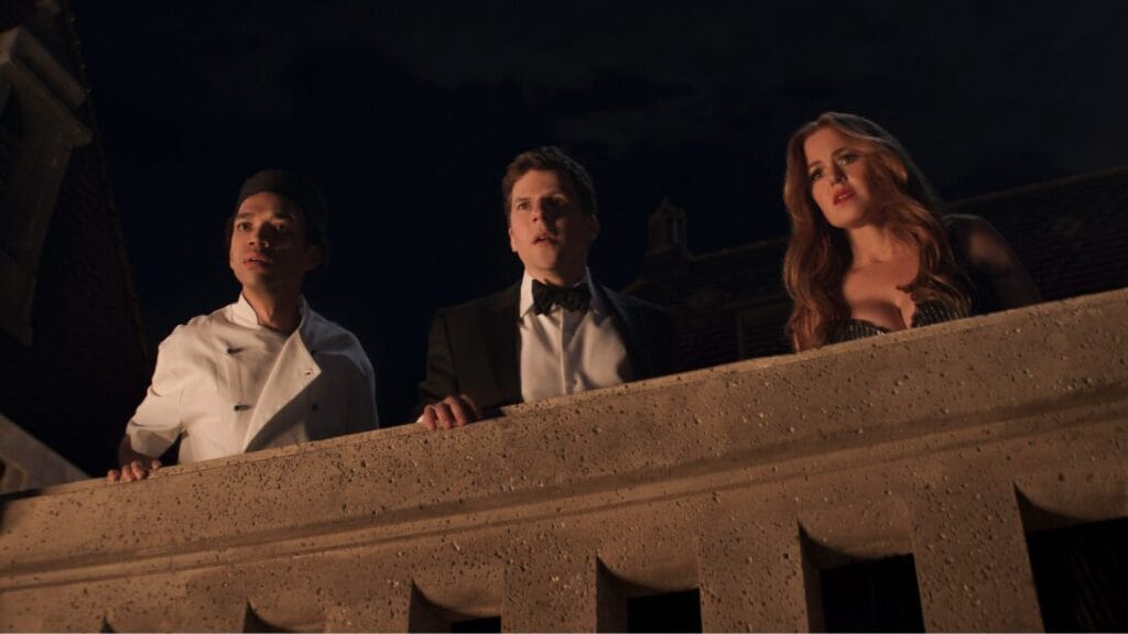 Justice Smith, Jesse Eisenberg, and Isla Fisher look down at a fancy party from a dark rooftop while wearing luxurious suits and dresses in NOW YOU SEE ME 3. 