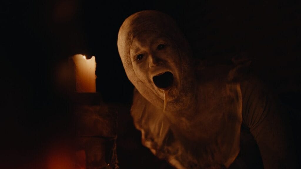 A horrifying pale-faced humanoid monster with wax dripping out of its mouth crawls next to a lit candle in a dark room in KEEPER directed by Osgood Perkins.